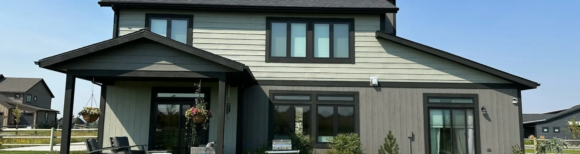 Front view of a modern gray two-story house with large windows, a covered patio, and hanging flower baskets on a green lawn under a clear blue sky.