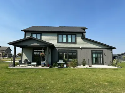 Front view of a modern gray two-story house with large windows, a covered patio, and hanging flower baskets on a green lawn under a clear blue sky.