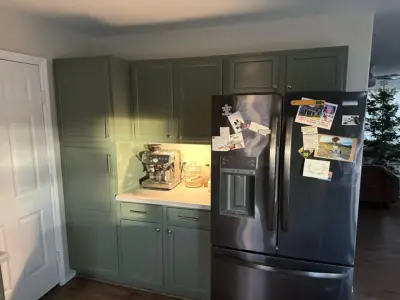 Kitchen with sage-green cabinets and a stainless fridge covered in magnets and photos; espresso machine on the counter.