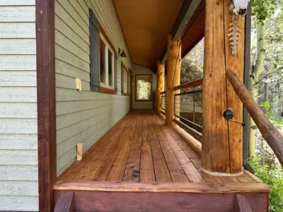 Rustic cabin-style porch with thick wooden log columns, a wooden deck, and light green siding with red trim along windows and doors, leading to an interior doorway. A bone-like ornament hangs on one post near the railing.