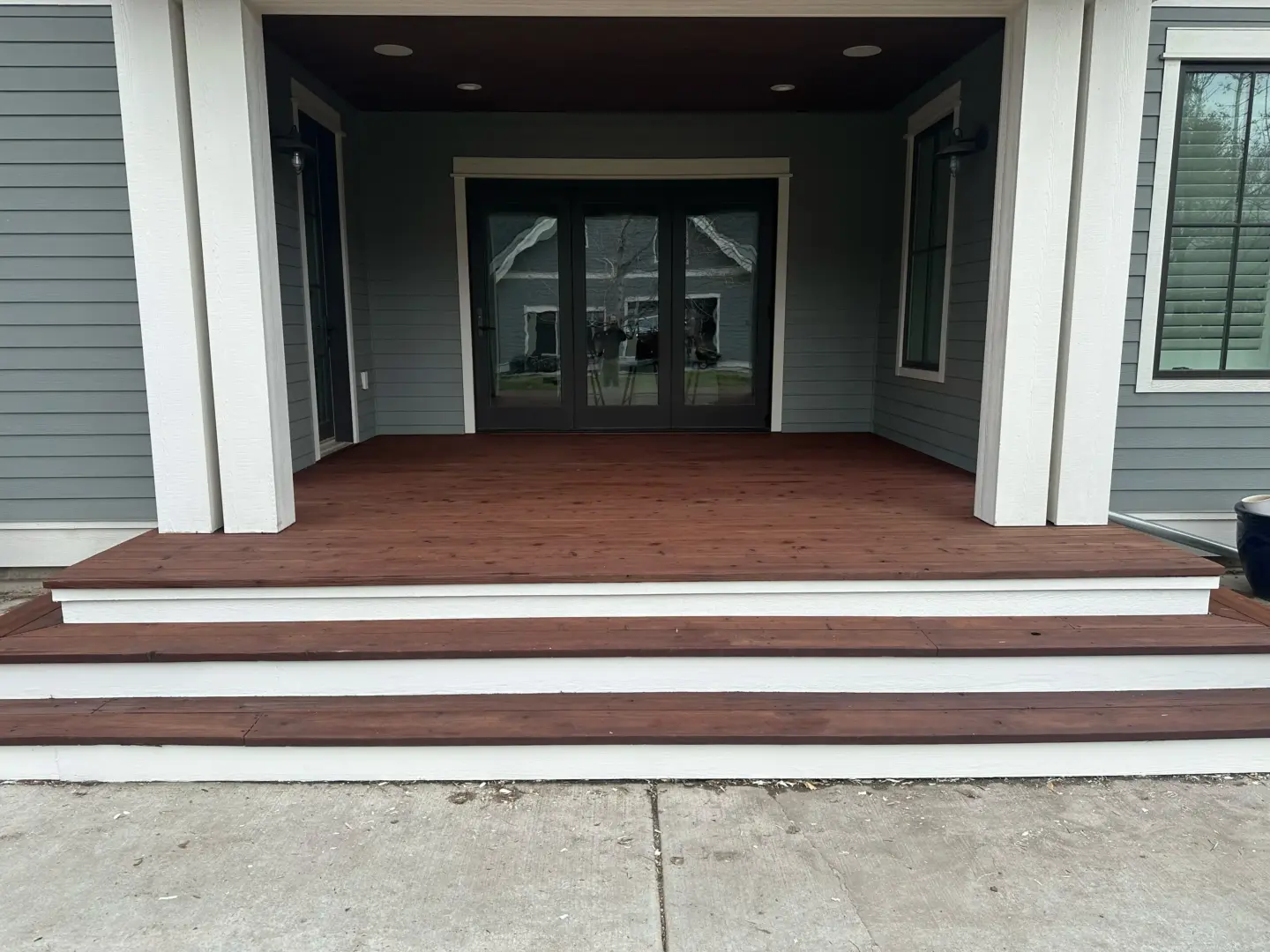 Front porch entry with brown wooden deck, white trim columns, and black double doors with glass panes reflecting the yard and trees behind.