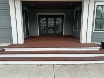 Front porch entry with brown wooden deck, white trim columns, and black double doors with glass panes reflecting the yard and trees behind.