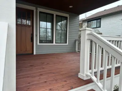 Front porch with brown door and large framed windows, gray siding, and white railing on a wooden deck.