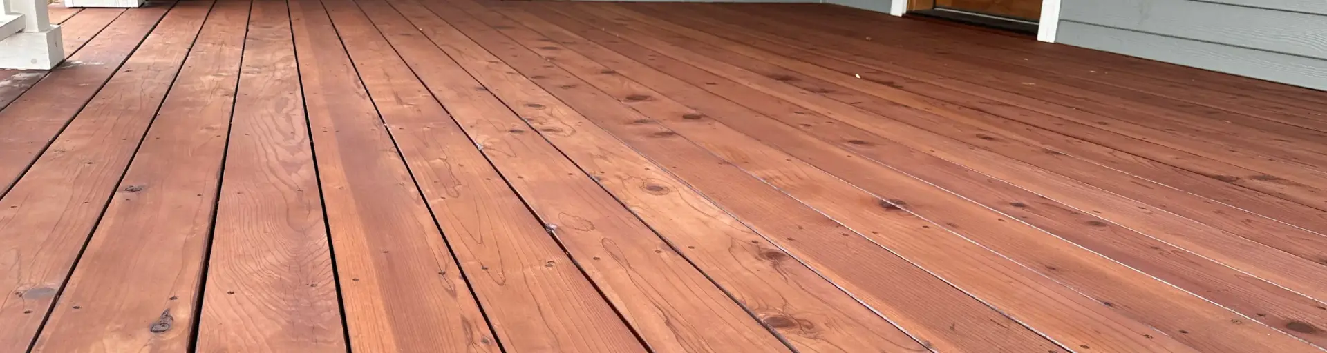 Wooden front porch/deck with reddish-brown planks in front of a gray-siding house, white window trim, and a wooden entry door on the right.