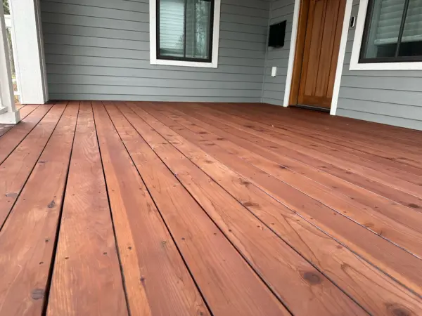 Wooden front porch/deck with reddish-brown planks in front of a gray-siding house, white window trim, and a wooden entry door on the right.