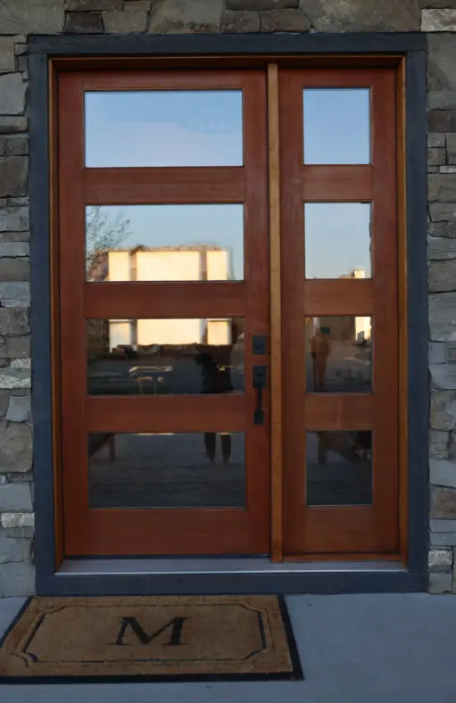 Double wooden front door with glass panes, set in a stone-framed entryway, with a brown doormat outside.