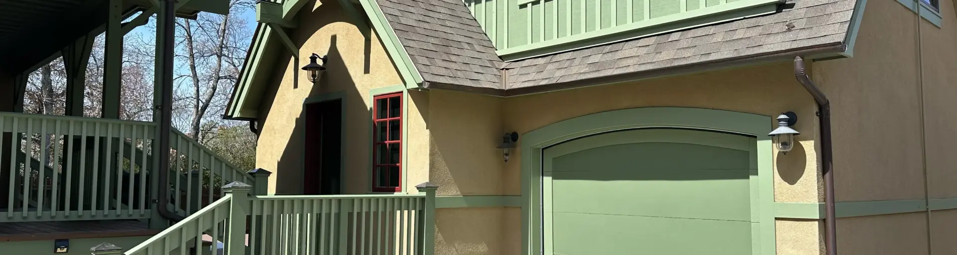 Exterior of a house with green trim, beige walls, and a green garage door under a steep roof.