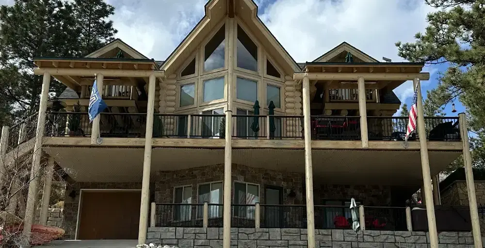 Log-style two-story house with a wide balcony supported by pillars, stone lower level, and flags on the railing.