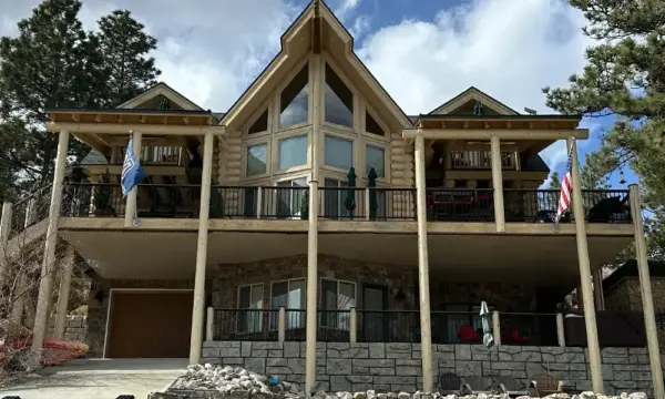 Log-style two-story house with a wide balcony supported by pillars, stone lower level, and flags on the railing.