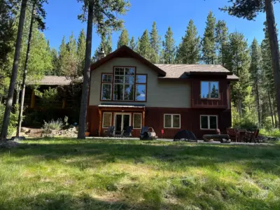 Two-story wooden house with red trim in a pine forest, blue sky above, and a patio with chairs outside.