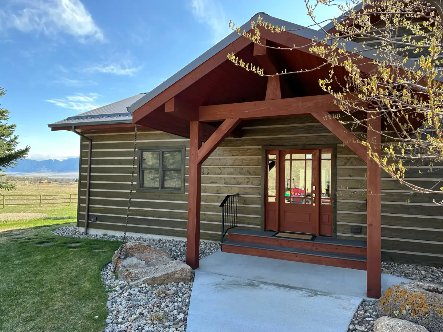 Front porch of a rustic house with red wooden beams, glass-paned door, and gravel path; mountains visible in the distance under a blue sky.