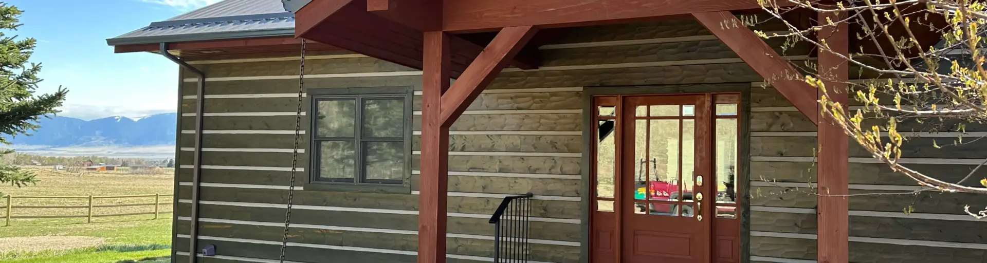 Front porch of a rustic house with red wooden beams, glass-paned door, and gravel path; mountains visible in the distance under a blue sky.