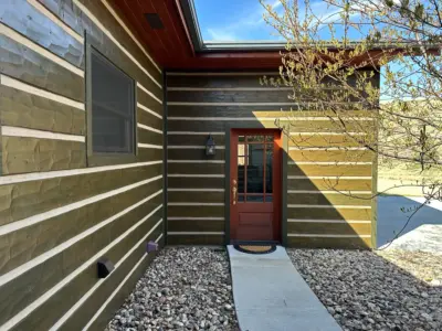 Front wooden house with a red door, gray stepping stones, and a gravel path bordered by a leafless tree on the right.