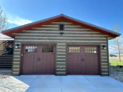 Front view of a two-car garage with horizontal wood siding, red double doors, and a peaked roof against a clear blue sky.