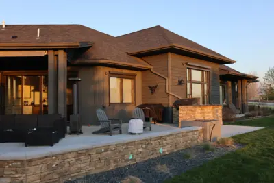 Backyard patio adjacent to a modern house with a stone retaining wall, outdoor seating, and a barbecue grill in evening light
