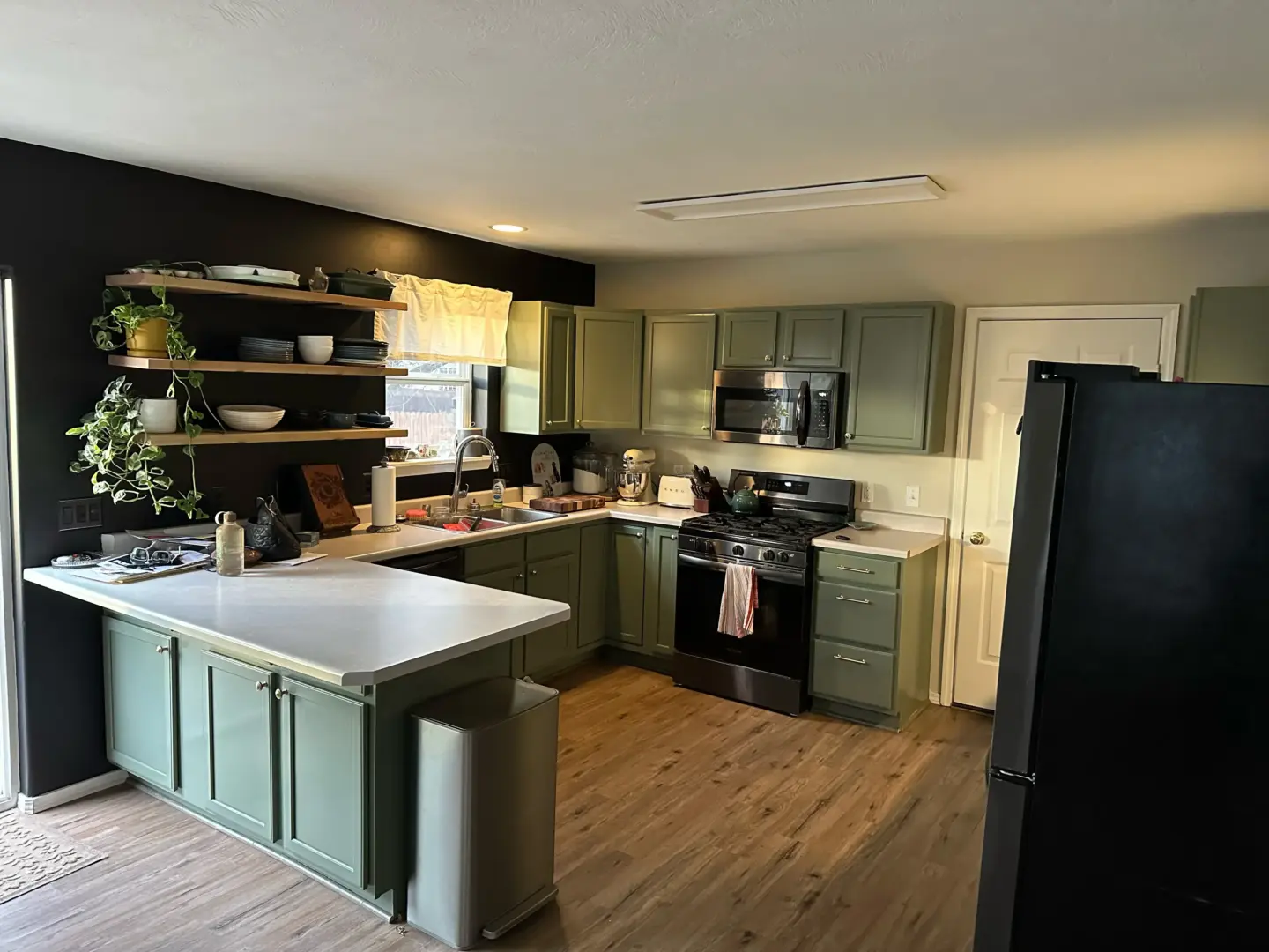 Green kitchen with mint cabinets, white countertops, and open wood shelves holding dishes and plants.