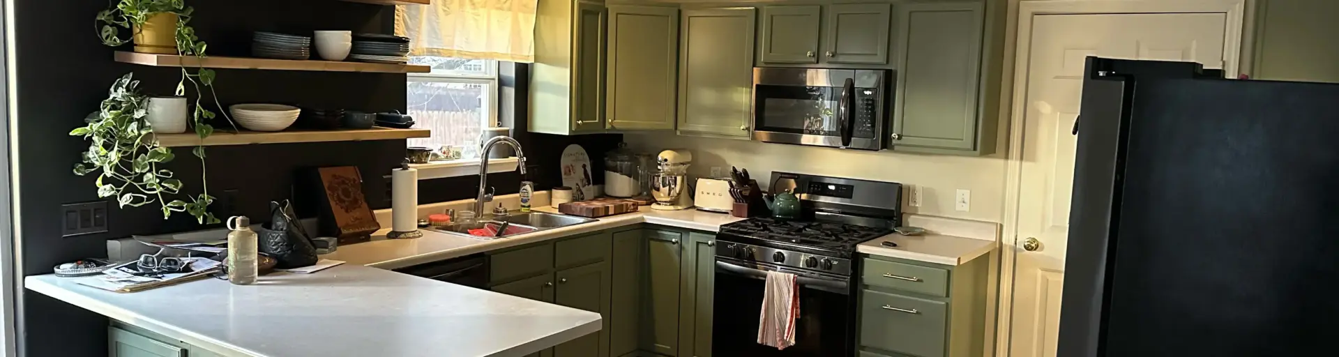 Green kitchen with mint cabinets, white countertops, and open wood shelves holding dishes and plants.