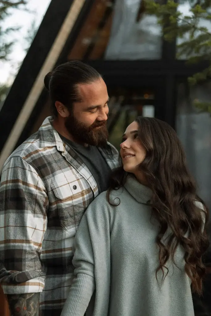 Bearded man in a plaid shirt gazes lovingly at a woman in a light gray sweater as they smile at each other outdoors.