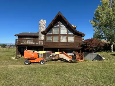 Brown wooden house with a triangular glass-front under renovation; plastic sheeting covers the windows and a tall stone chimney stands to the left, with an orange construction lift in the yard.