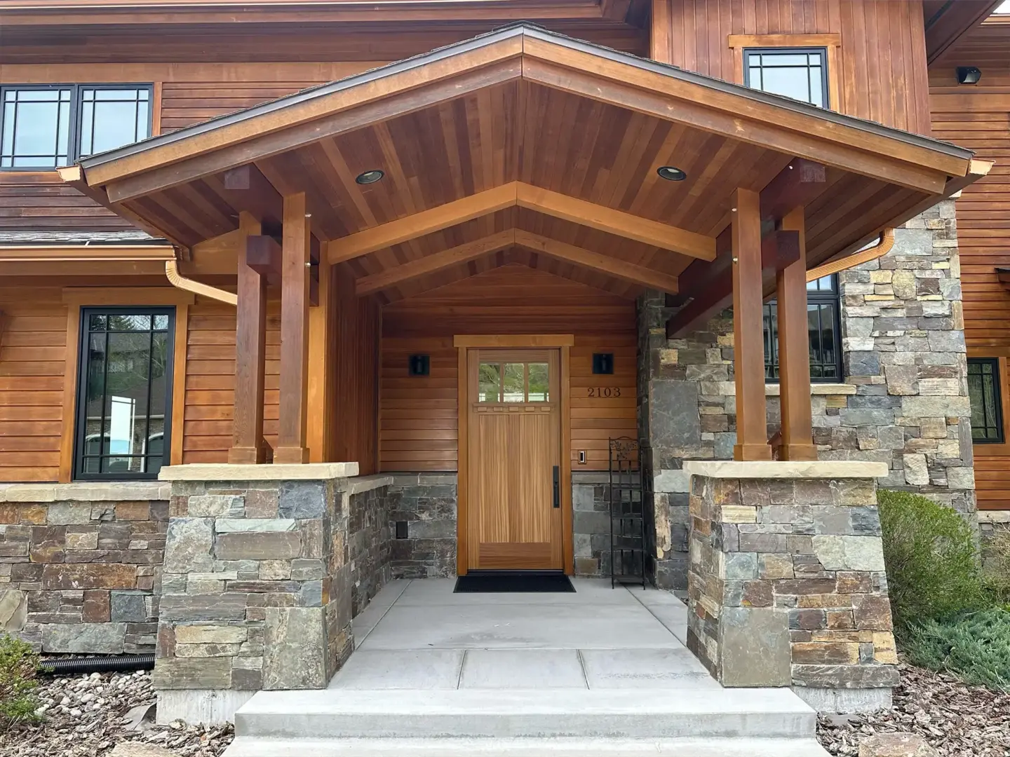 Wooden house entrance with a peaked cedar porch, stone columns, and a wooden door marked 2103.