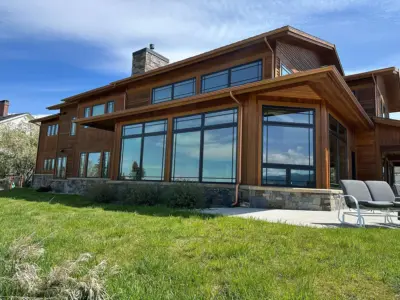 Front view of a modern wooden house with large glass windows, stone chimney, and a green lawn under a blue sky.