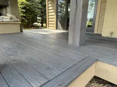 Wooden deck with gray planks and an outdoor cushioned seating area beside a fireplace, next to a sliding glass door into the house.