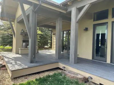 Wooden deck attached to a house, with large weathered posts supporting a covered patio and sliding glass doors nearby.